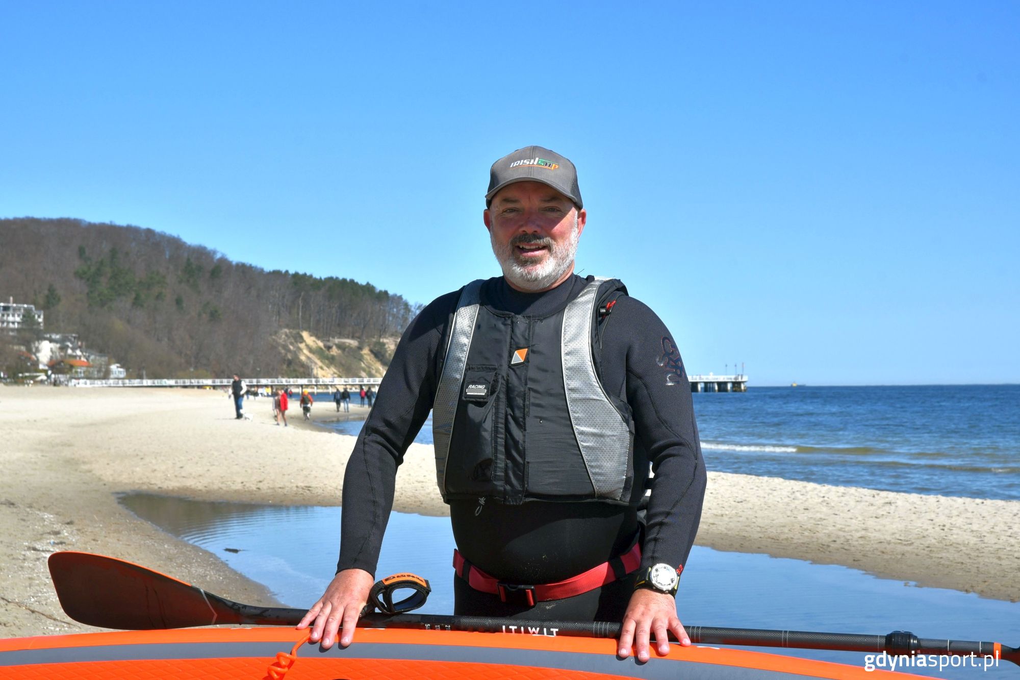 Paul Byrne w piance z deska i wiosłem i deską na tle molo
